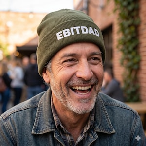 May include: An olive green beanie with the word "EBITDAD" in white block letters. The beanie is worn by a person wearing a denim jacket and a plaid shirt. The person is smiling.
