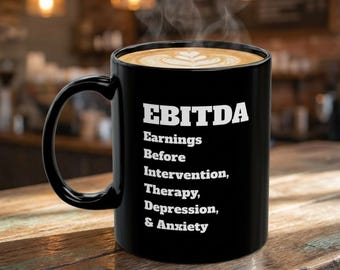 May include: A black ceramic mug filled with coffee featuring white text that reads EBITDA Earnings Before Intervention, Therapy, Depression, & Anxiety. The mug has a curved handle and is filled to the brim with a latte art design.