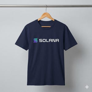 May include: Navy blue t-shirt with the word "SOLANA" in white block letters. The word is above a stylized logo in shades of blue and green. The t-shirt is hanging on a wooden hanger.
