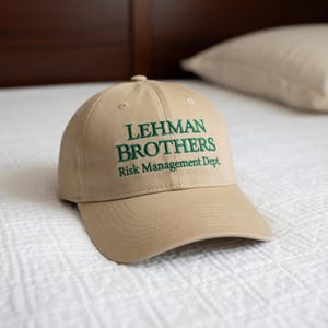 Op de afbeelding: Een beige baseballcap met de groen geborduurde tekst "LEHMAN BROTHERS Risk Management Dept.". De pet ligt op een wit oppervlak.