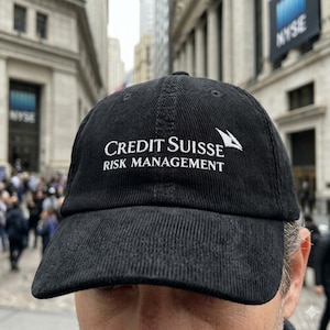 Può includere: Cappellino da baseball in velluto a coste nero con la scritta bianca "CREDIT SUISSE RISK MANAGEMENT". Il cappellino ha una visiera curva e una corona strutturata. Lo sfondo mostra una strada cittadina con l'insegna NYSE.