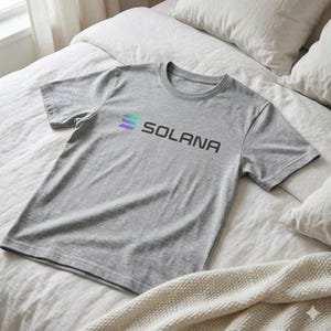 May include: A gray t-shirt with the word "SOLANA" printed in black, accompanied by a stylized logo in shades of green, blue, and purple. The shirt is made of a soft material and is displayed on a bed.
