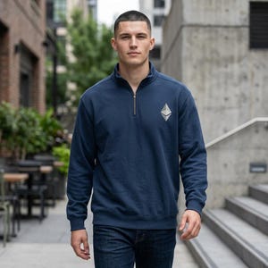Puede incluir: Una sudadera azul marino con cremallera de cuarto de círculo y un logotipo plateado de Ethereum en el pecho izquierdo. La sudadera tiene un cuello alzado y una cremallera plateada. La persona lleva vaqueros azules.