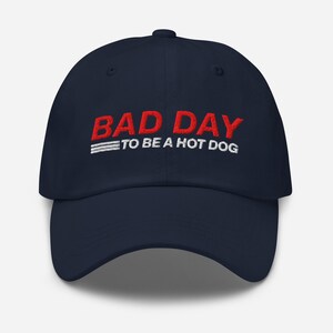 以下が含まれることがあります： 紺色の野球帽。赤い文字で「BAD DAY」、その下に白い文字で「TO BE A HOT DOG」と書かれています。帽子はカーブしたつばと、布製のストラップ留めが付いています。