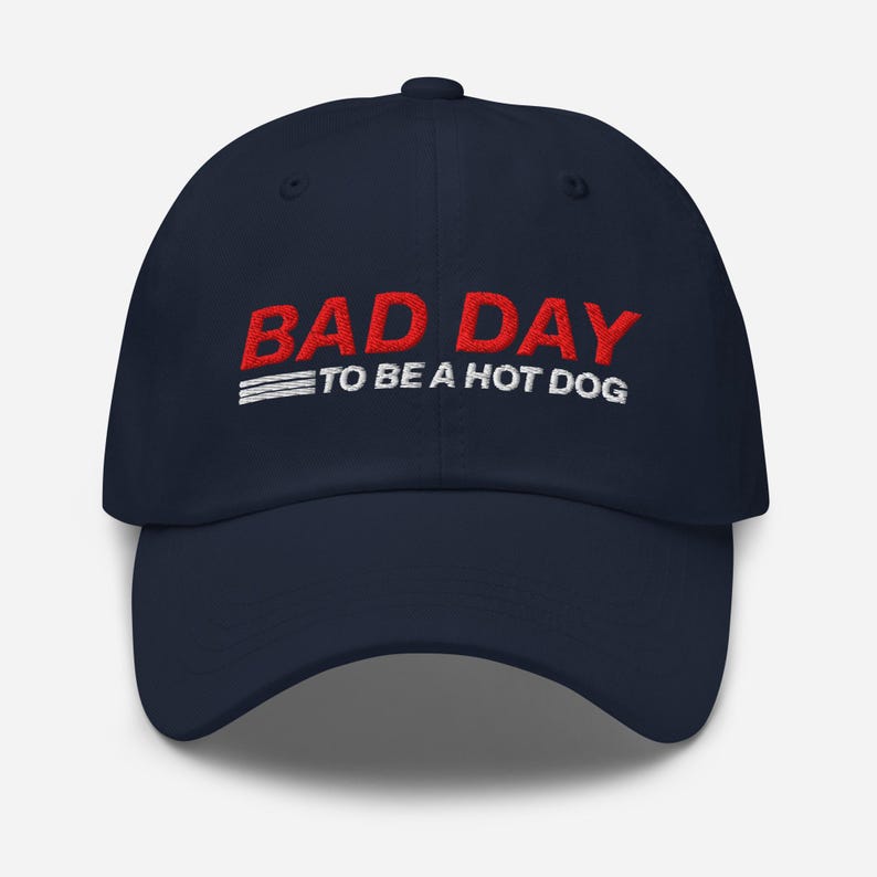 以下が含まれることがあります： 紺色の野球帽。赤い文字で「BAD DAY」、その下に白い文字で「TO BE A HOT DOG」と書かれています。帽子はカーブしたつばと、布製のストラップ留めが付いています。