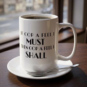 May include: A white ceramic coffee mug filled with dark coffee, resting on a white saucer with a silver spoon. The mug has the text "IF COP A FEEL I MUST THEN COP A FEEL I SHALL" in black lettering.