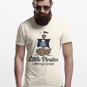 Könnte beinhalten: Cremefarbenes T-Shirt mit einem Cartoon-Piratenschiff mit Totenkopf und gekreuzten Knochen auf dem Segel. Der Text "Little Pirates Learning Center" ist darunter gedruckt. Das T-Shirt wird von einer Person getragen.