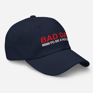 以下が含まれることがあります： 赤色の「BAD DAY」と白の「TO BE A HOT D」の文字が入った、ネイビーブルーの野球帽。帽子にはカーブしたつばと、上部にボタンが付いています。テキストは帽子の前面に刺繍されています。