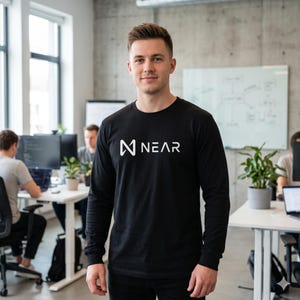 Op de afbeelding: Een zwart shirt met lange mouwen met de witte tekst "NEAR" en een gestileerd logo. Het shirt wordt gedragen door een persoon in een kantooromgeving. Andere mensen werken op de achtergrond aan bureaus.