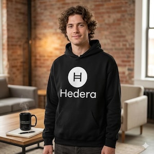 Può includere: Felpa con cappuccio nera con logo e testo Hedera bianchi. La felpa ha una tasca frontale e un cappuccio con coulisse. La persona nell'immagine indossa la felpa. Una tazza nera con testo è su un tavolo.