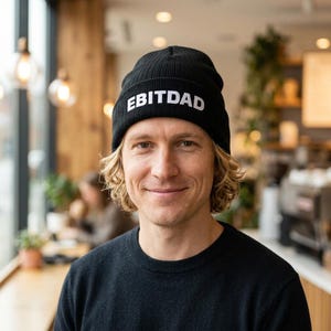 May include: A black beanie with the white text "EBITDAD" is worn by a person with blond hair. The person is wearing a black sweater. The background is a cafe setting with wooden accents and soft lighting.