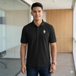Può includere: Polo nero con logo Ethereum argentato sul petto. La maglietta ha un colletto classico e maniche corte. La persona indossa jeans blu e un orologio marrone.