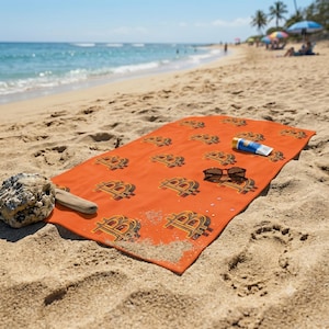 May include: A rectangular orange beach towel featuring a repeating pattern of black and gold Bitcoin symbols. A pair of dark-framed sunglasses and a small tube of sunscreen are placed on the towel, along with a piece of driftwood and a rock.