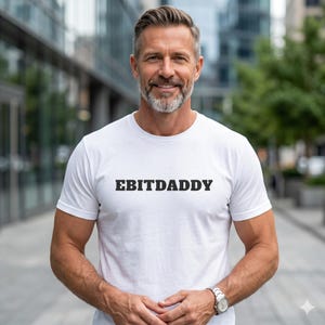 Könnte beinhalten: Weißes T-Shirt mit dem schwarzen Text "EBITDADDY". Eine Person trägt das T-Shirt und eine silberne Uhr. Der Hintergrund ist eine verschwommene Außenszene mit Gebäuden.
