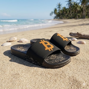Puede incluir: Sandalias negras con un símbolo dorado de Bitcoin en la parte superior, descansando en una playa arenosa. Las sandalias están cerca del océano, con conchas marinas y palmeras al fondo. Las sandalias están cubiertas de arena.