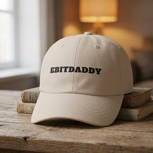 May include: A beige baseball cap featuring the text EBITDADDY embroidered in black thread across the front panel. The cap has a structured crown, a curved brim with visible stitching, and a button at the top centre of the fabric dome.