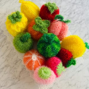 May include: A collection of colorful knitted fruit-shaped dishcloths in a white lace-edged bowl. The dishcloths are made to look like lemons, oranges, strawberries, peaches, and watermelons.