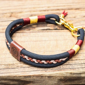 Puede incluir: Un collar para perros negro y marr&oacute;n con herrajes dorados. El collar presenta un dise&ntilde;o trenzado con detalles en rojo y beige. Una etiqueta de cuero marr&oacute;n con un logotipo est&aacute; adjunta.