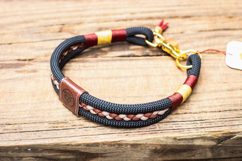 Puede incluir: Un collar para perros negro y marr&oacute;n con herrajes dorados. El collar presenta un dise&ntilde;o trenzado con detalles en rojo y beige. Una etiqueta de cuero marr&oacute;n con un logotipo est&aacute; adjunta.