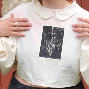 May include: A white cropped blouse with puffed sleeves and a peter pan collar. The blouse features a black and white print of a sword in a pool of water with stars.