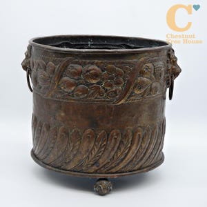 May include: A bronze-coloured planter with ornate floral and fruit carvings. The planter has a wide, cylindrical shape with lion head handles and rests on three decorative feet. The text "Chestnut Tree House" is visible in the upper right corner.