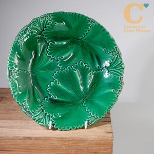 May include: A round, green ceramic plate with a leaf design. The plate has a glossy finish and is supported by white stands. The leaf pattern is detailed and textured, creating a three-dimensional effect. The plate is displayed on a wooden surface.
