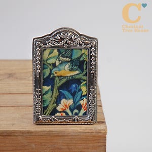May include: A rectangular silver-toned metal frame with an arched top and intricate embossed detailing. The frame contains a fabric insert featuring a colourful needlepoint design of a bird perched on a branch surrounded by green leaves and flowers.