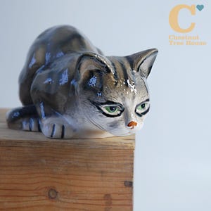 Könnte beinhalten: Eine Keramik-Katzenfigur in geduckter Haltung. Die Katze hat graue und braune Streifen, grüne Augen und eine kleine orangefarbene Nase. Die Figur liegt auf einer Holzoberfläche. Das Logo von Chestnut Tree House ist im Hintergrund.