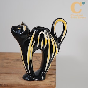 May include: A black ceramic cat-shaped pitcher with yellow and white stripes. The pitcher has a handle and a spout, and the cat is standing on its hind legs. The pitcher is on a wooden surface.