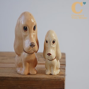 Könnte beinhalten: Zwei Keramik-Hundefiguren. Der größere Hund ist hellbraun, der kleinere cremefarben. Beide haben braune Augen und Nasen, Schlappohren und ein Halsband. Sie sitzen auf einer Holzoberfläche. Die Figuren sind dekorativ.