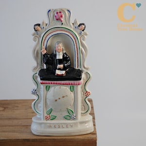 Könnte beinhalten: Vintage Keramikfigur eines Predigers in schwarzer Robe, stehend hinter einem schwarzen Pult. Die Figur ist weiß mit farbigen Akzenten, darunter rote, grüne und blaue Details. Das Wort "WESLEY" ist auf der Basis aufgedruckt.