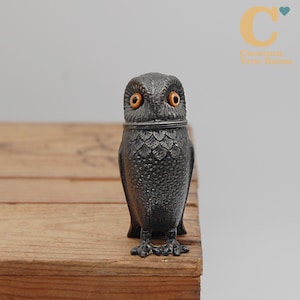 May include: A dark gray owl figurine with orange eyes. The owl has a textured surface and is perched on a wooden surface. The figurine has a hinged head and is likely a decorative item.