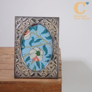 May include: A silver-toned picture frame with an oval aperture, displaying a floral print on a light blue backdrop. The frame features an elaborate, embossed design with floral and ribbon patterns. The frame is placed on a wooden surface.