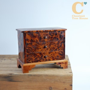 Può includere: Un piccolo comò antico in legno con finitura lucida e fantasia. Il comò ha tre cassetti con pomelli scuri e poggia su una base in legno coordinata. La venatura del legno è visibile e il colore generale è marrone caldo. Il testo "Chestnut Tree House" è nell'angolo in alto a destra.