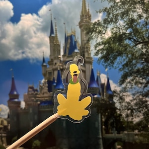 May include: A yellow Pluto character sticker on a wooden stick, with a green collar and a black outline. The background features a blurred image of a castle and a blue sky with clouds. The sticker is transparent.