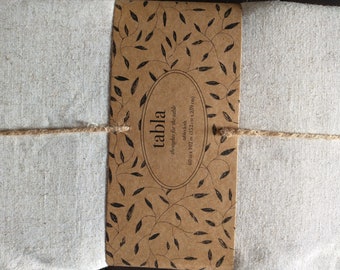 May include: A folded, off-white tablecloth tied with twine. A brown paper label is wrapped around the cloth, featuring the word "tabla" in a circular design, along with the text "thoughts for the table" and dimensions of 60 x 102 inches (152 cm x 259 cm).