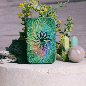May include: A small, green tin with a swirling, colorful design featuring a stylized lotus flower. The tin is surrounded by small yellow flowers and green leaves.