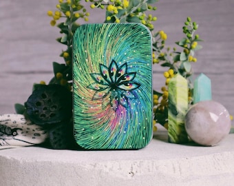 May include: A small, green tin with a swirling, colorful design featuring a stylized lotus flower. The tin is surrounded by small yellow flowers and green leaves.