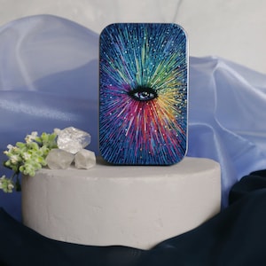 Op de afbeelding: Een klein, rechthoekig blikje met een kleurrijk, abstract ontwerp met een groot oog in het midden. Het ontwerp doet denken aan een sterrenstelsel met een regenboog van kleuren die vanuit het oog naar buiten stralen.