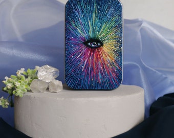 May include: A small, rectangular tin with a colorful, abstract design featuring a large eye in the center. The design is reminiscent of a galaxy with a rainbow of colors radiating outward from the eye.