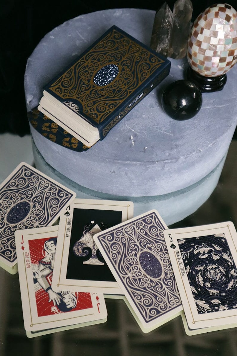 K&ouml;nnte beinhalten: Ein Kartenspiel mit einem dunkelblauen und goldenen Design. Die Karten zeigen einen sternenklaren Nachthimmel und komplizierte Muster. Das Deck hei&szlig;t "Natural Magic".