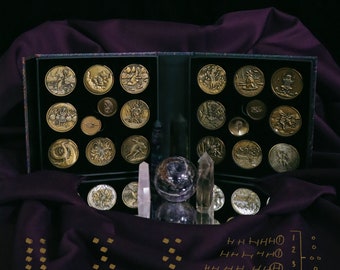 May include: Gold-coloured coins with engraved designs are displayed in a black box. The box is open, revealing multiple coins. Crystals, a purple sphere, and a purple cloth are also present in the image.