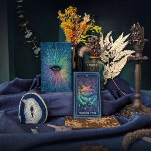 May include: A tarot card with a blue background and a green frog with a rainbow pattern. The card is titled "Prismatic Toad" and is surrounded by dried flowers and a blue cloth.