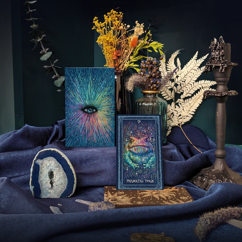 May include: A tarot card with a blue background and a green frog with a rainbow pattern. The card is titled "Prismatic Toad" and is surrounded by dried flowers and a blue cloth.