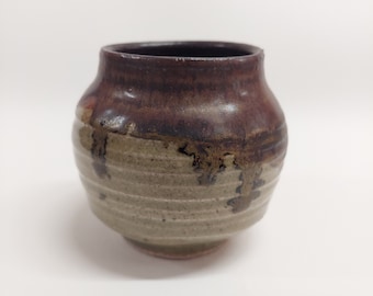 Könnte beinhalten: Eine handgefertigte Keramikvase mit rundem Körper und dunkelbraunem Rand. Die Vase hat ein strukturiertes, erdiges Aussehen mit einem hellbraunen Sockel und dunkleren braunen Tropfen. Die Keramik ist wahrscheinlich auf der Töpferscheibe gedreht und hat eine rustikale, organische Ästhetik.