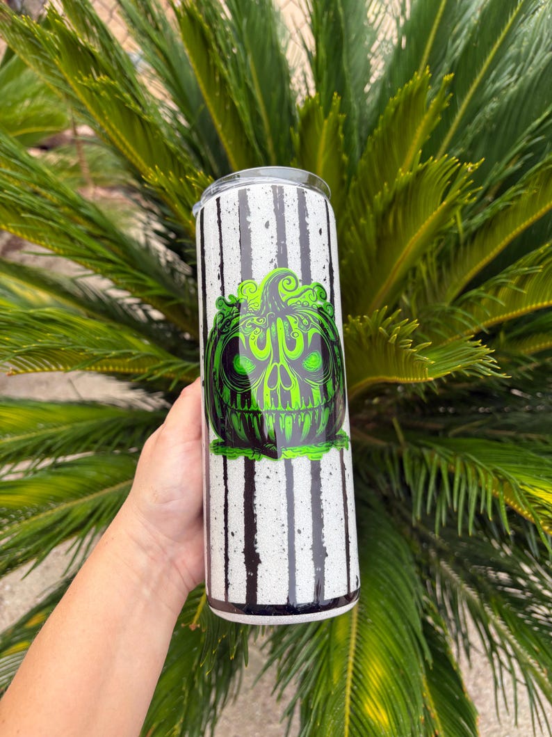 May include: A tall, stainless steel tumbler with a black and white striped pattern. The tumbler features a green and black graphic of a skull-like pumpkin. The design is likely for Halloween or a similar theme.