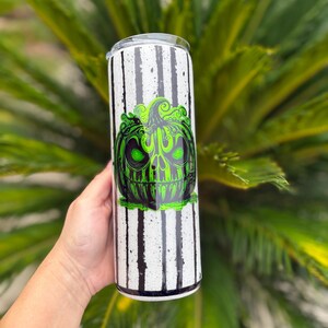 May include: A white tumbler with black vertical stripes and a green and black pumpkin design. The pumpkin has a skull-like face. The tumbler is held in a person's hand.