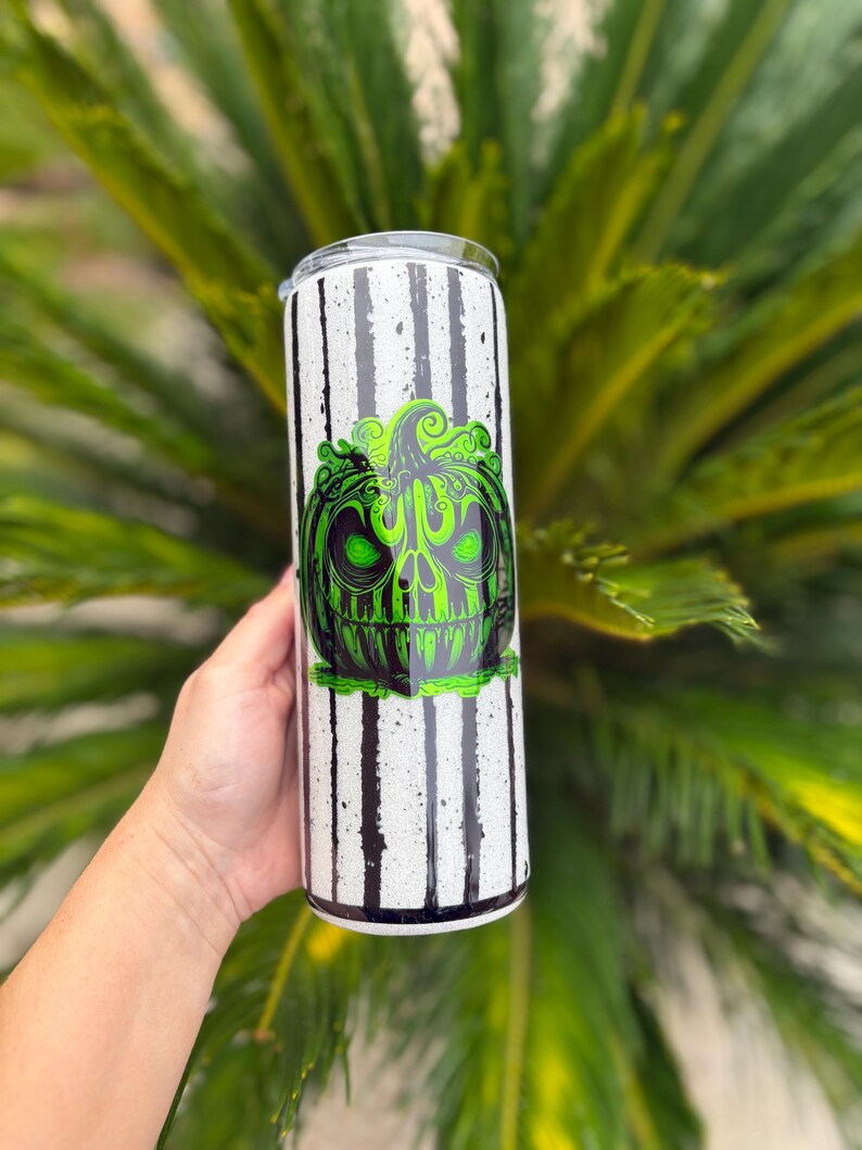 May include: A white tumbler with black vertical stripes and a green and black pumpkin design. The pumpkin has a skull-like face. The tumbler is held in a person's hand.