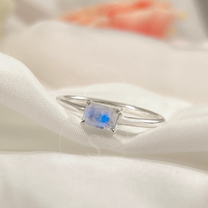 May include: A silver ring featuring a rectangular moonstone with an iridescent sheen. The ring sits on white silk fabric, with blurred flowers in the background. The ring has a slender band with a split design near the moonstone.