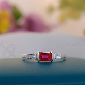 Op de afbeelding: Een zilveren ring met een rechthoekige, levendige rode edelsteen als middelpunt, geflankeerd door twee kleinere, witte baguette-geslepen stenen. De ring ligt op een teal oppervlak, met een zachte, vage bloemenachtergrond.